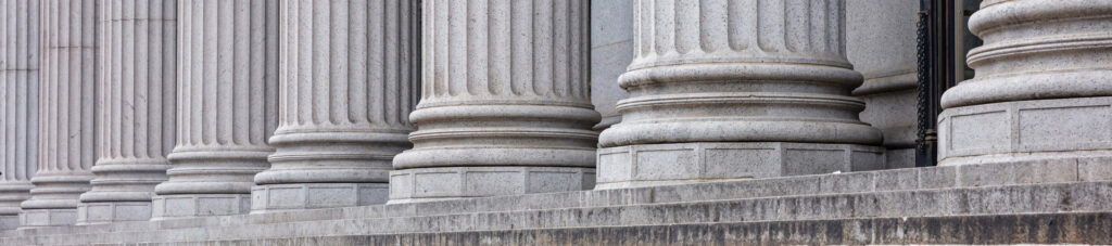A row of large, gray stone columns with fluted shafts and detailed bases stands atop a stone staircase, suggesting the exterior of a courthouse